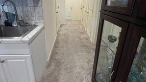 A narrow hallway with a kitchen area on the left, a dark wooden cabinet with glass doors on the right, and a carpeted floor.