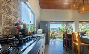 A kitchen with a gas stove, a refrigerator, and a dining area with wooden chairs and a table. - Beautiful House With Pool, Arraial D'ajuda Lvl001 (Porto Seguro)