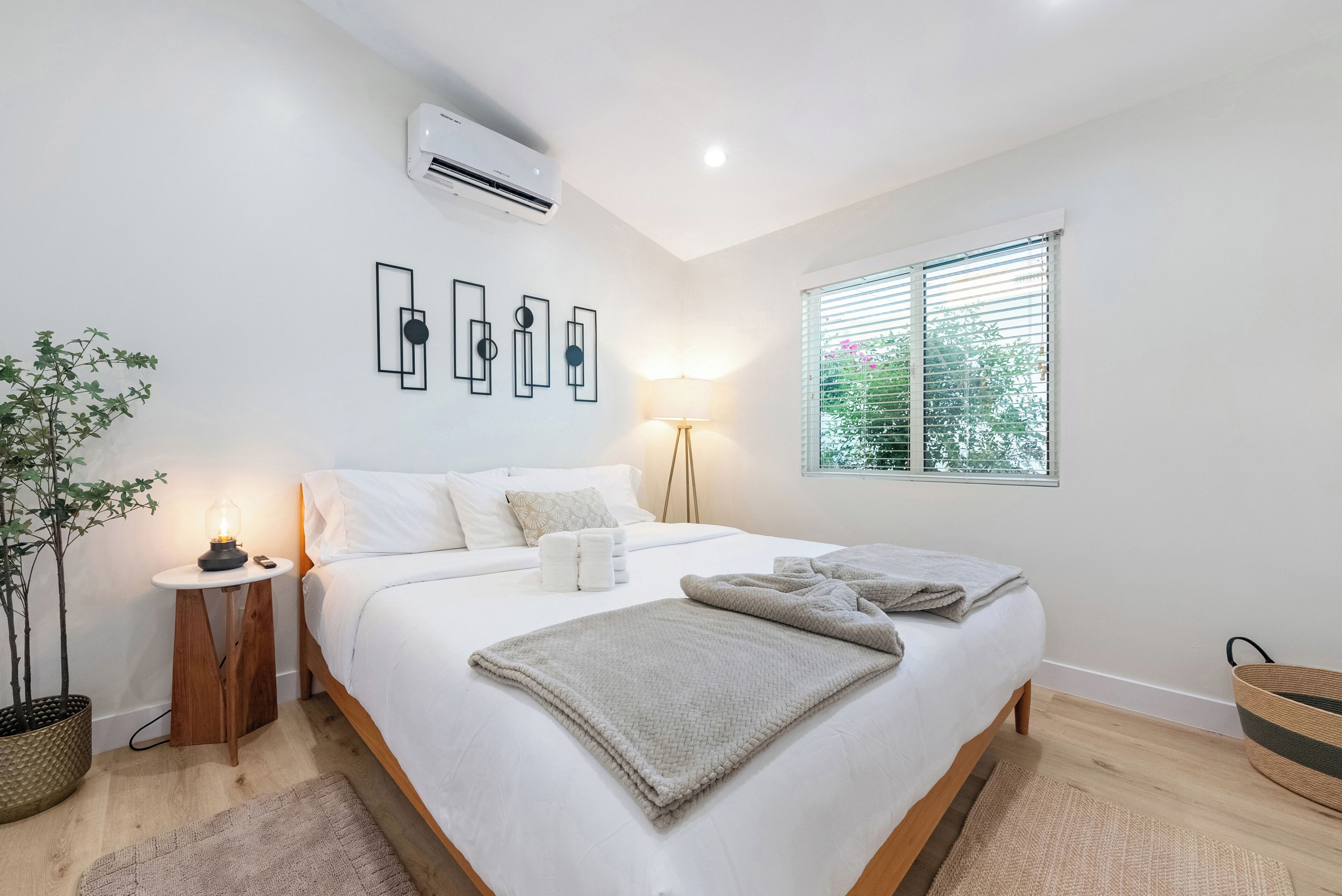 Un dormitorio con cama, mesita de noche, lámpara, aire acondicionado, decoración en la pared, ventana con persianas y planta en maceta.