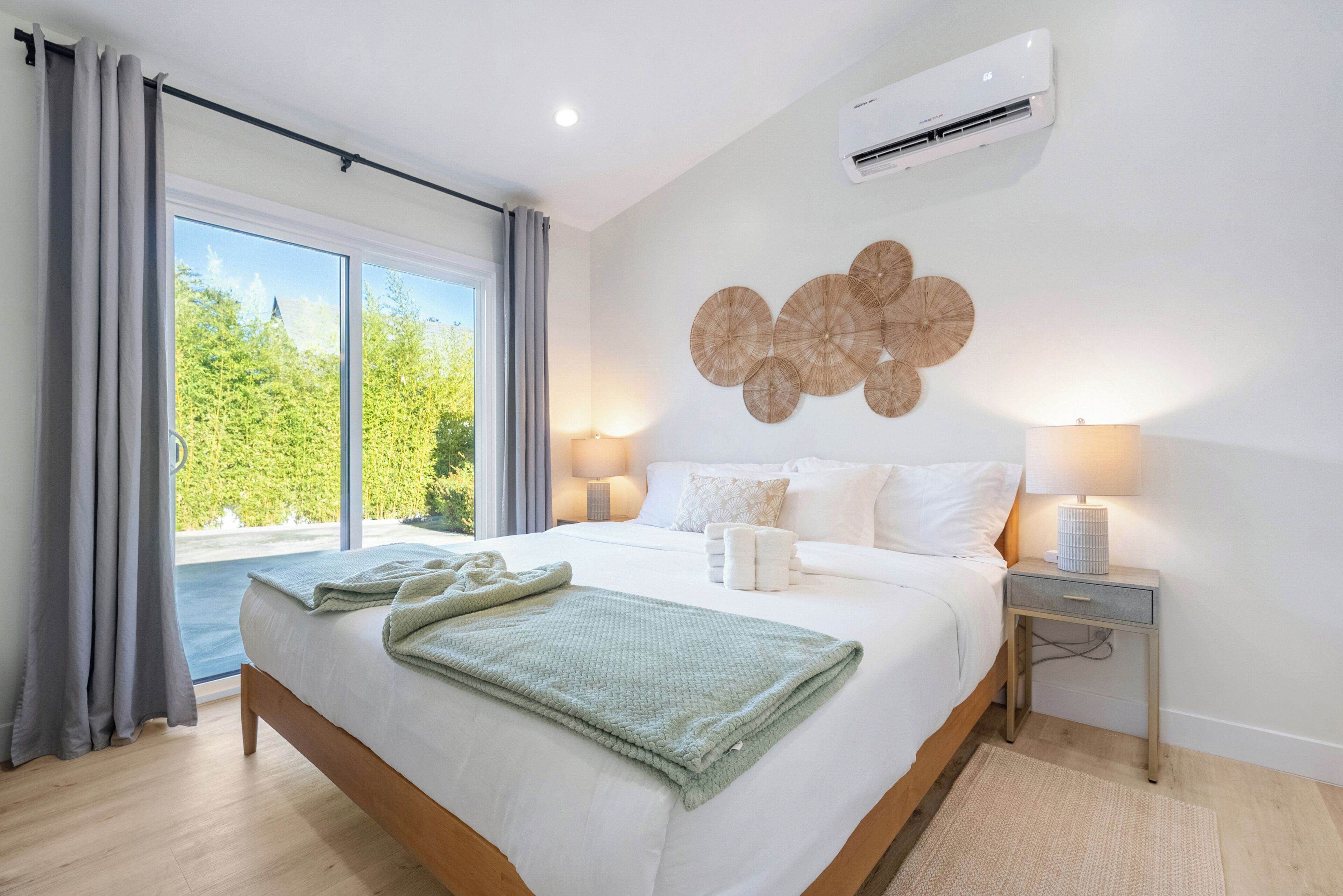 A hotel room with a large bed, two bedside lamps, a wall-mounted air conditioner, and a view of greenery outside.