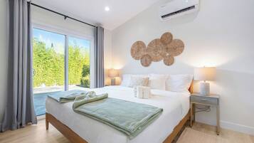 A hotel room with a large bed, two bedside lamps, a wall-mounted air conditioner, and a view of greenery outside.