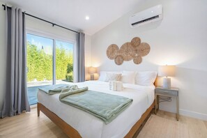A hotel room with a large bed, two bedside lamps, a wall-mounted air conditioner, and a view of greenery outside. - Modern 2BR Pasadena Guesthouse w/ King Bed & Outdoor Kitchen & AC (Pasadena)