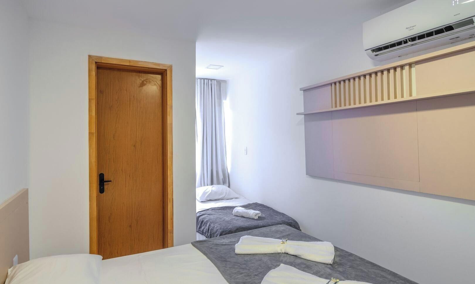 Quarto de hotel com porta de madeira, cama com manta cinzenta, toalha sobre a cama, janela com cortinas e ar condicionado fixado na parede.