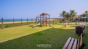 Un parque infantil con un tobogán y estructuras para escalar, rodeado por una valla, con una playa y el océano al fondo.