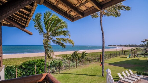 Vista a la playa desde un espacio cubierto con terraza de madera, palmeras y un parque infantil.