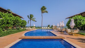 Zona de piscina con sillas de descanso y sombrillas, rodeada de vegetación y palmeras.