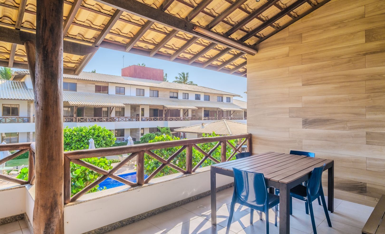 Un balcón con una mesa y sillas de madera, vista a un edificio con techo de tejas y una piscina al fondo.