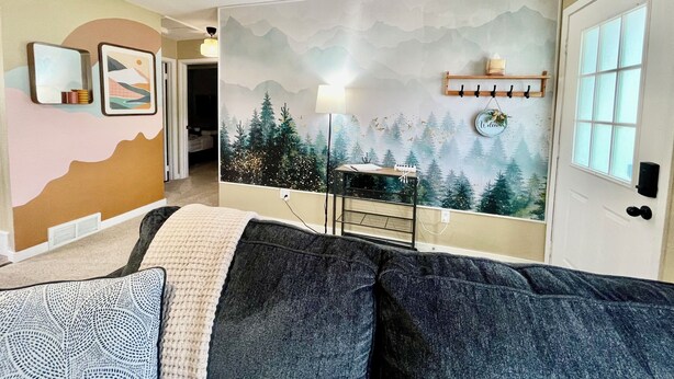 Ein gemütliches Wohnzimmer mit einer großen Couch, einer Wandmalerei von einem Wald, einer Lampe und einem kleinen Tisch.