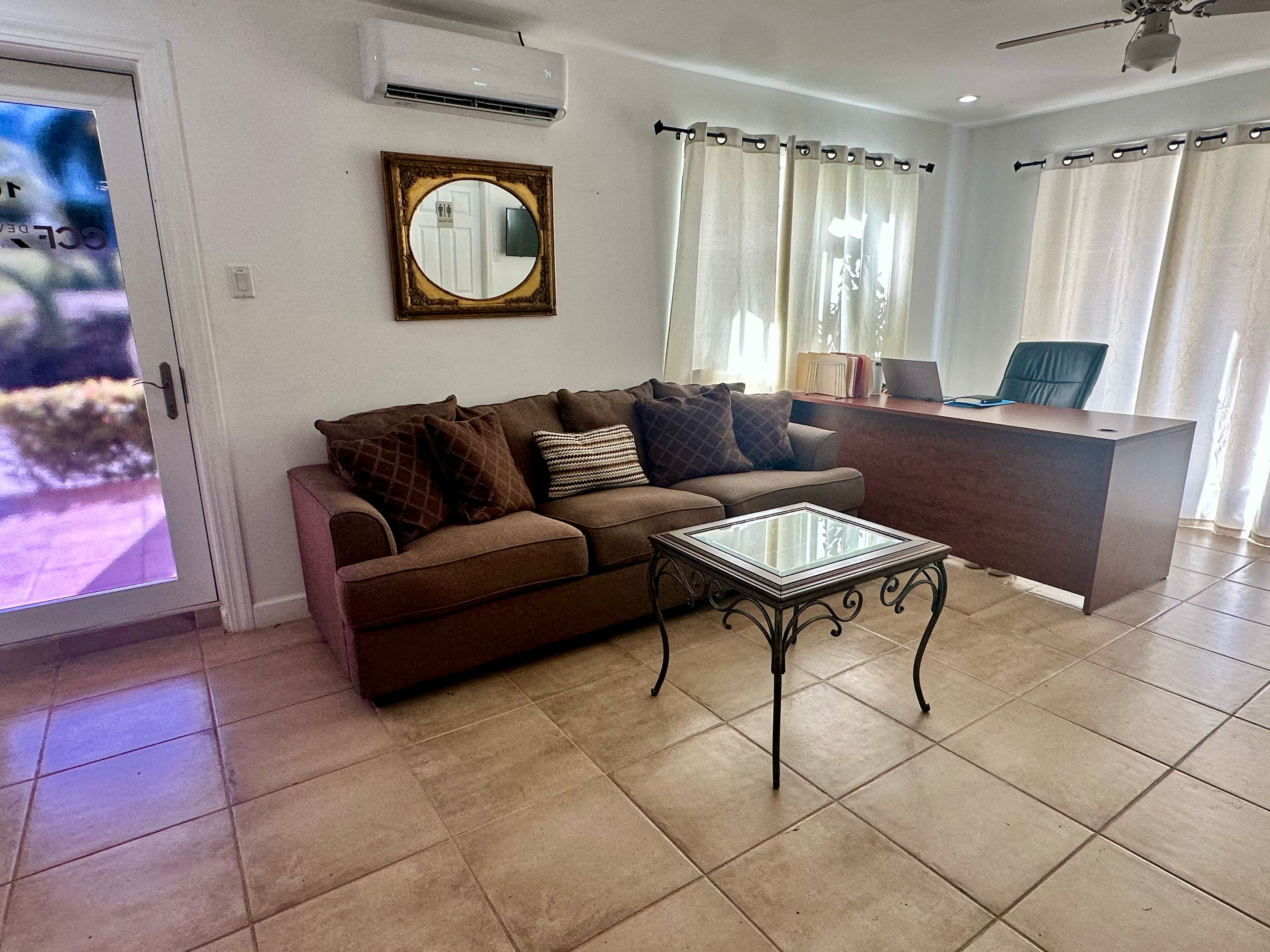A living room with a brown sofa, a glass coffee table, a desk with a chair, and a ceiling fan.