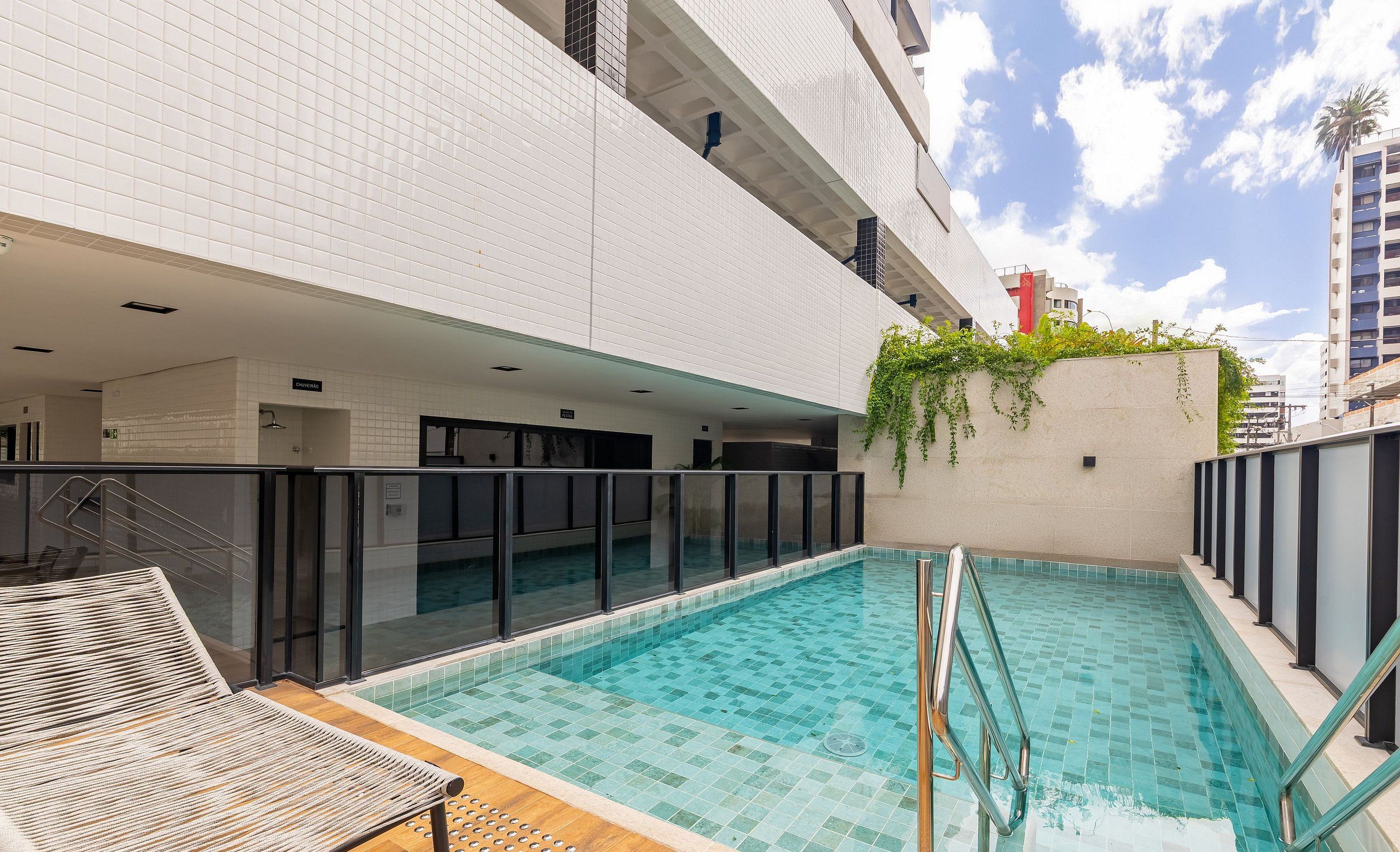 Una piscina interior con cubierta de madera y sillones de descanso, rodeada por un edificio moderno con paredes de azulejos y barandilla de vidrio.