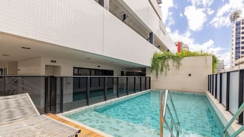 Una piscina interior con cubierta de madera y sillones de descanso, rodeada por un edificio moderno con paredes de azulejos y barandilla de vidrio.