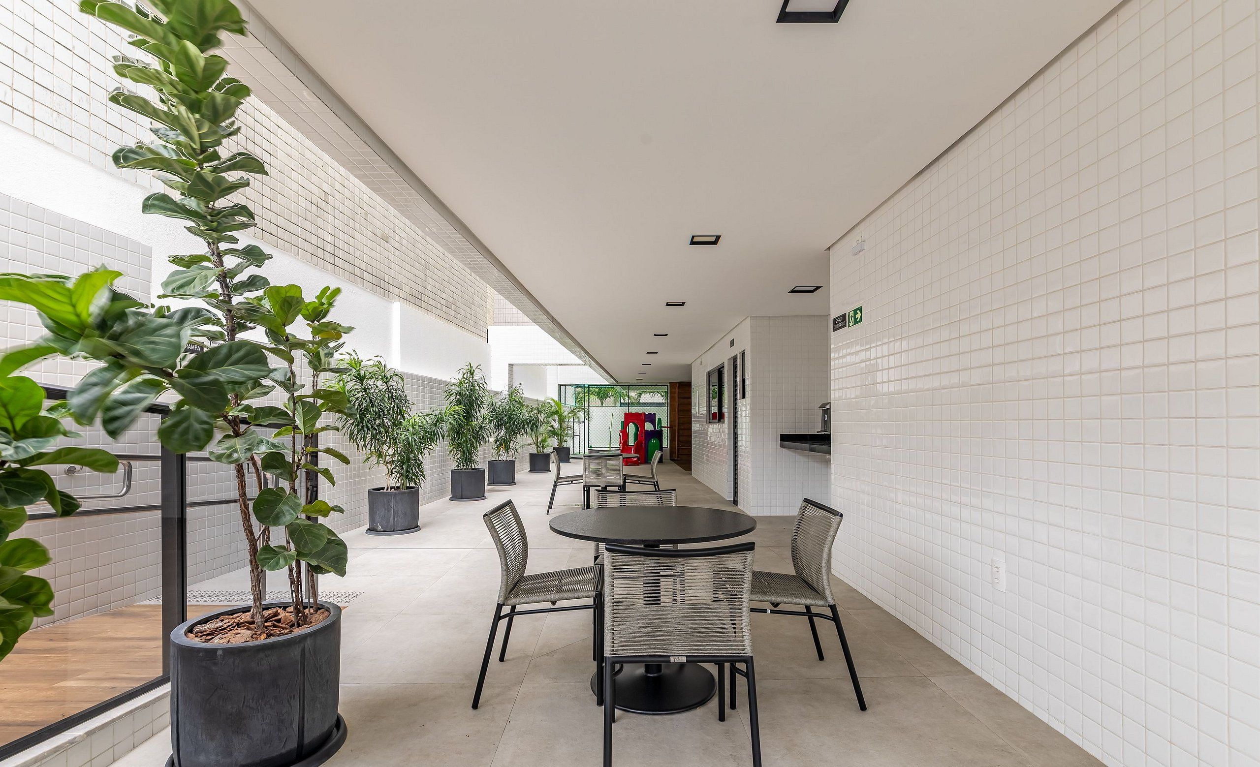 Un espacio interior moderno con un largo pasillo, plantas en macetas, una mesa redonda con sillas y una zona de juegos al fondo.