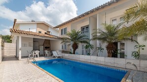 Una casa de dos pisos con piscina, mobiliario exterior y una chimenea de ladrillo.