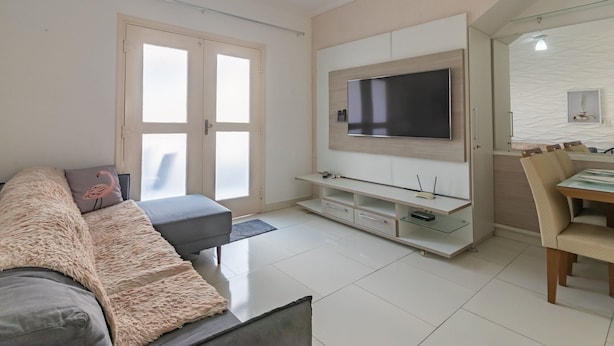 Una sala de estar moderna con un televisor de pantalla plana montado en la pared, un sofá gris con una manta suave, y un comedor con mesa y sillas.