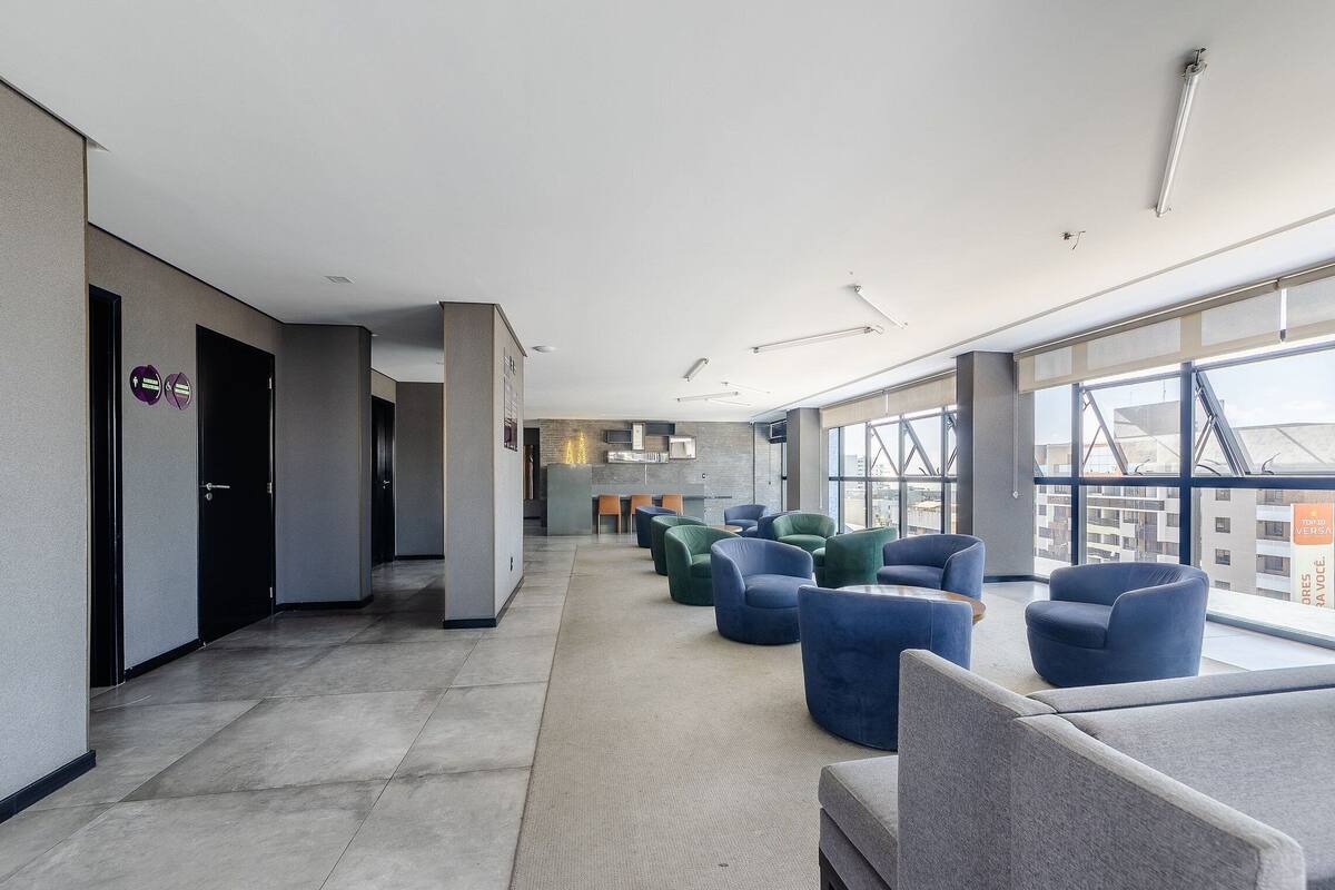A modern hotel lobby with a long corridor, seating area with blue and green chairs, and large windows offering city views.