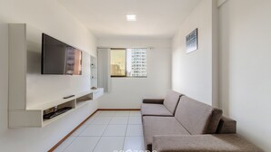 A modern living room with a flat-screen TV mounted on a white wall, a sofa, and a small window with a view of buildings. - Apart w/ Pool 450m From the Beach, Maceio Eez0301 (Maceió)