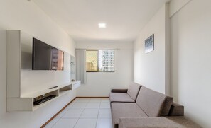 A modern living room with a flat-screen TV mounted on a white wall, a sofa, and a small window with a view of buildings. - Apart w/ Pool 450m From the Beach, Maceio Eez0301 (Maceió)