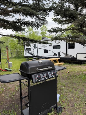 A portable gas grill with controls, a picnic table, and a recreational vehicle in the background. - Montana Moon on the Lake "Imagine" RV #6 (Babb)