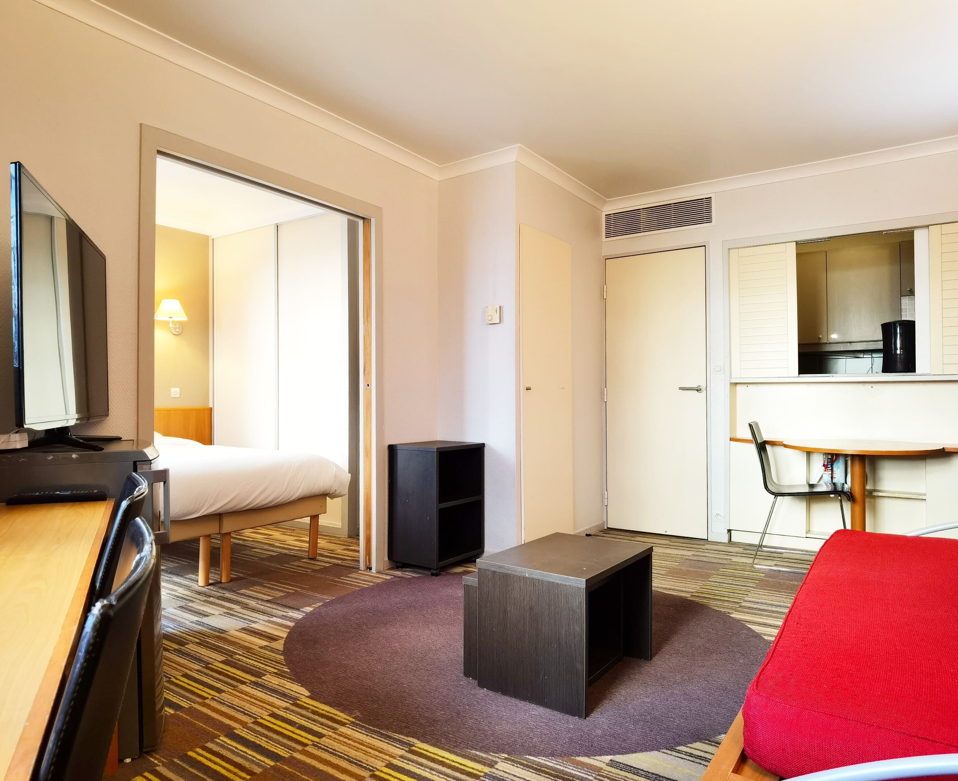 Une chambre d’hôtel comprenant un lit, un bureau avec une télévision, une petite table, une chaise rouge et un coin cuisine.