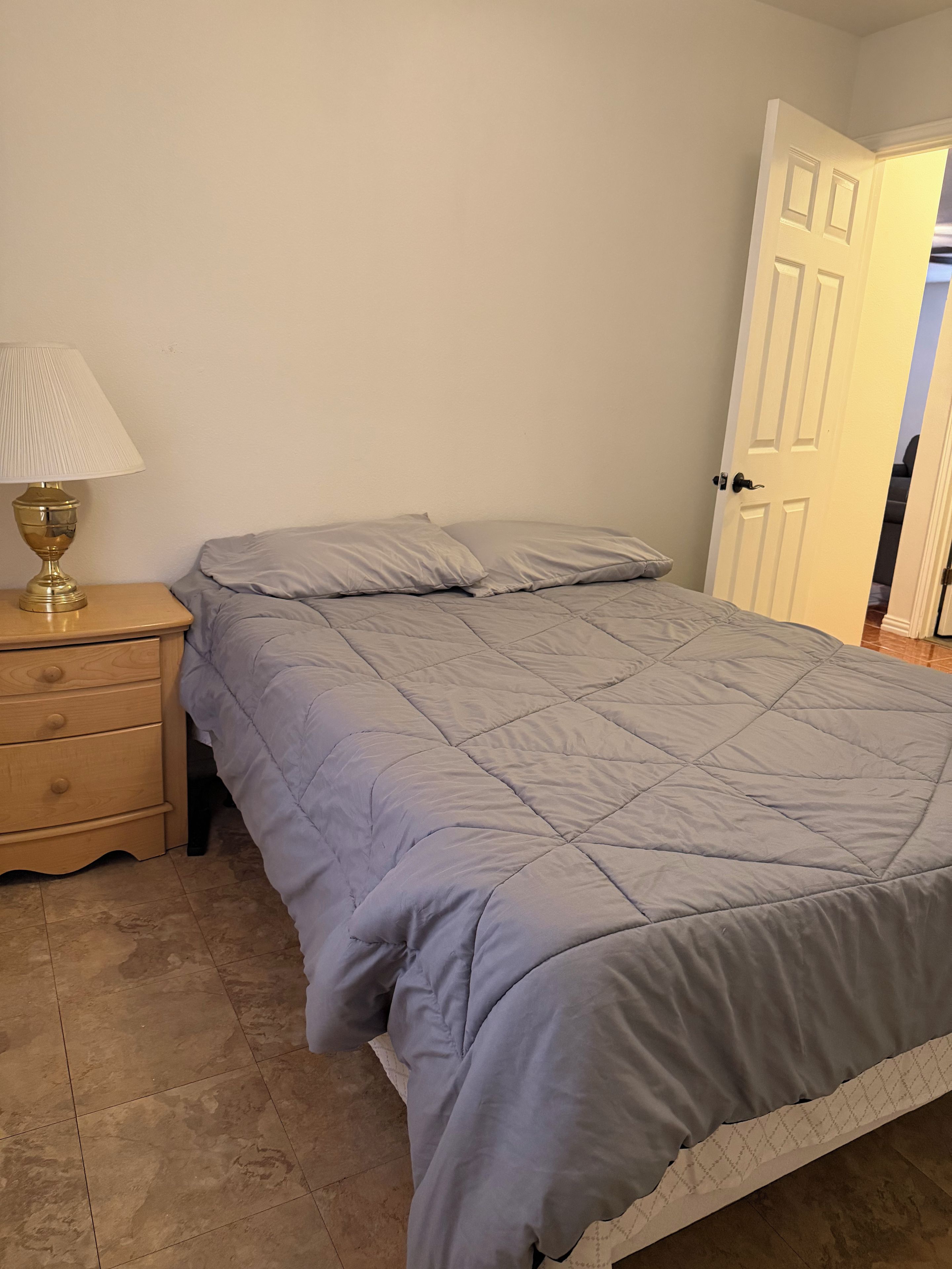 A bedroom with a bed, a nightstand with a lamp, and a door leading to another room.