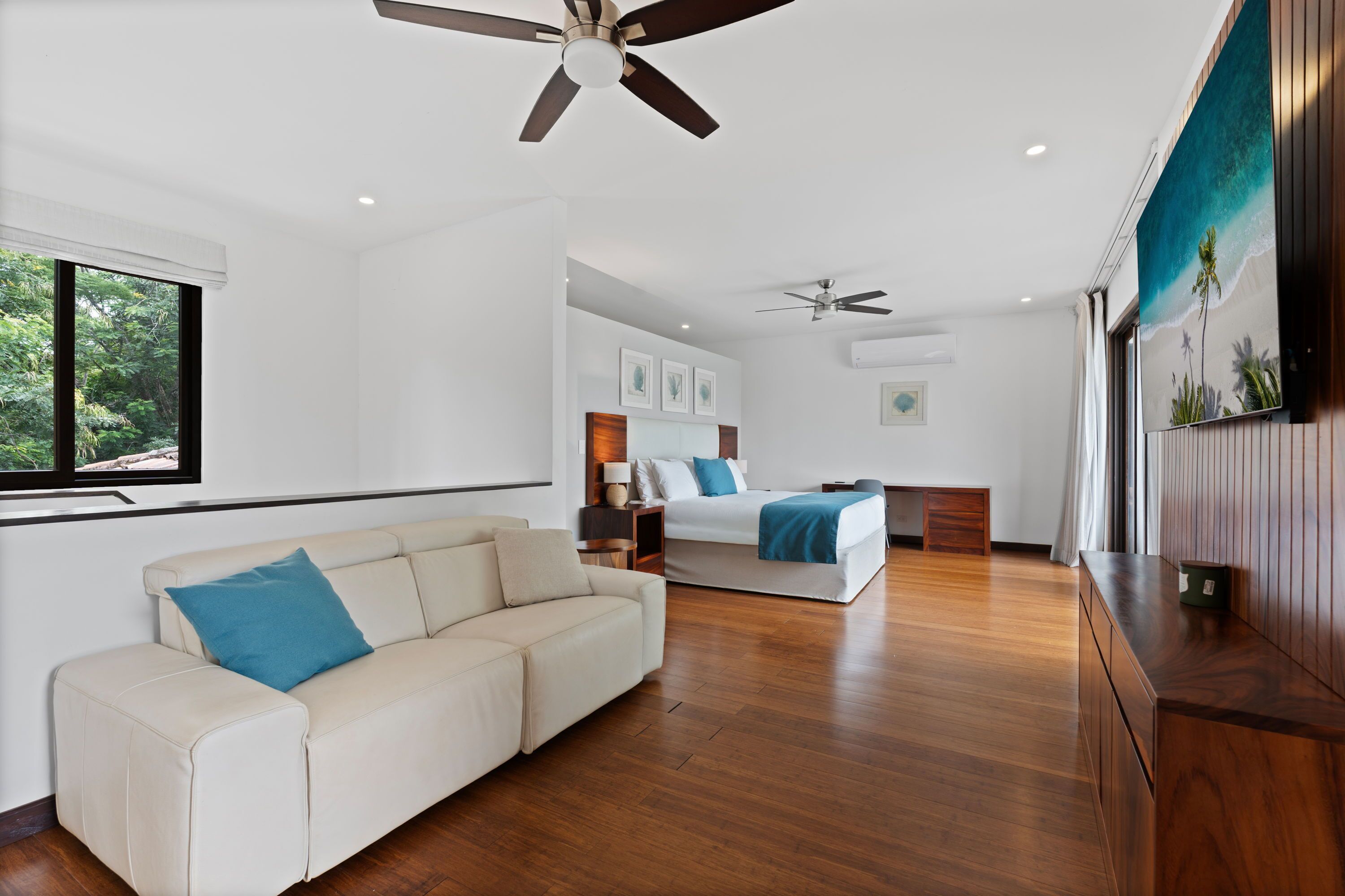 A spacious room with a wooden floor, a white sofa, a bed with a blue blanket, and a ceiling fan.
