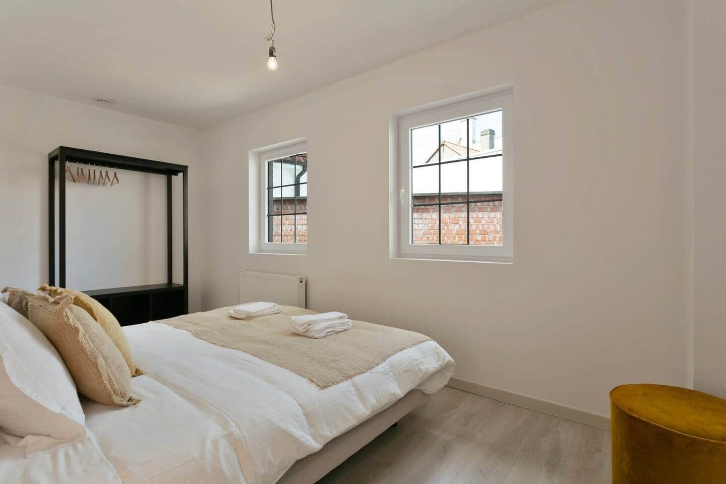 A bedroom with a bed, two windows, a radiator, and a small stool.