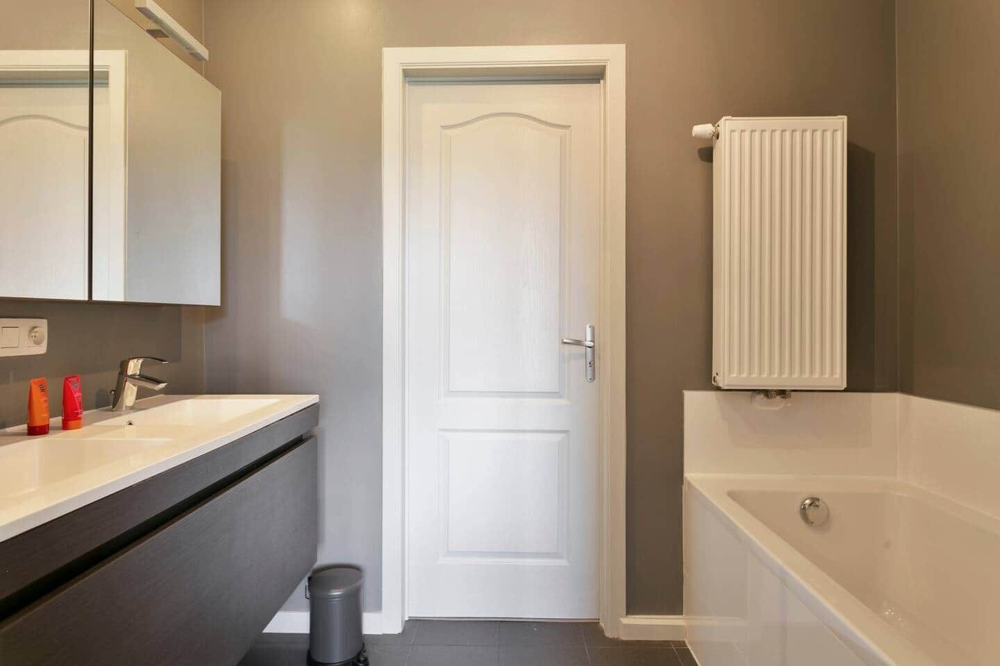 A bathroom with a white door, a bathtub, a white radiator, and a sink with two orange bottles.