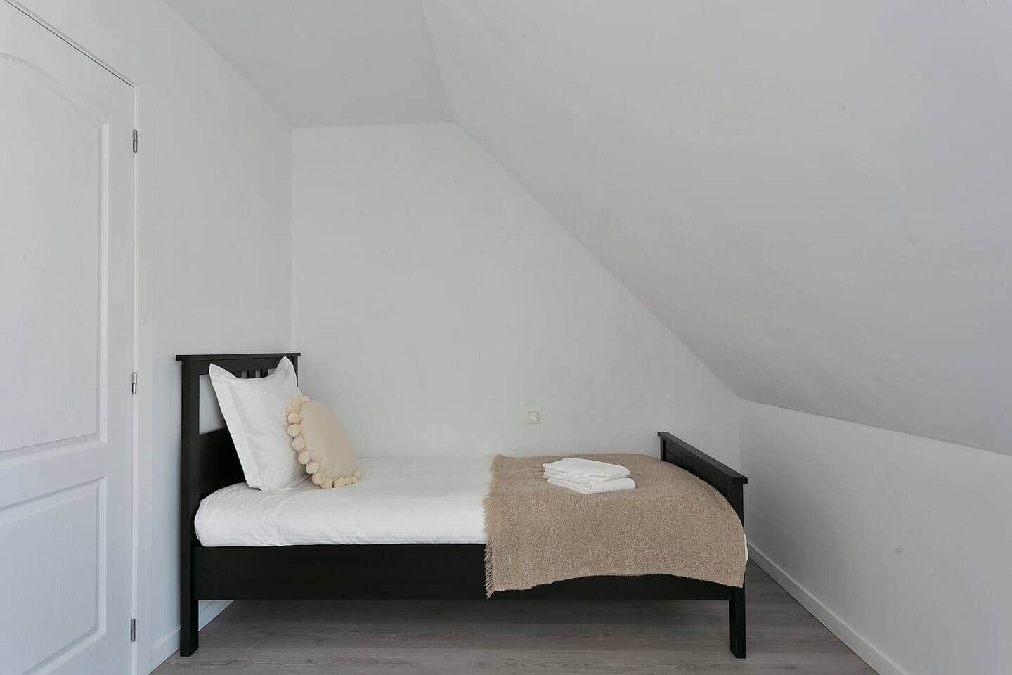 Une petite chambre avec un lit simple, un plafond en pente, un cadre de lit en bois foncé, du linge de lit blanc et un plaid beige.