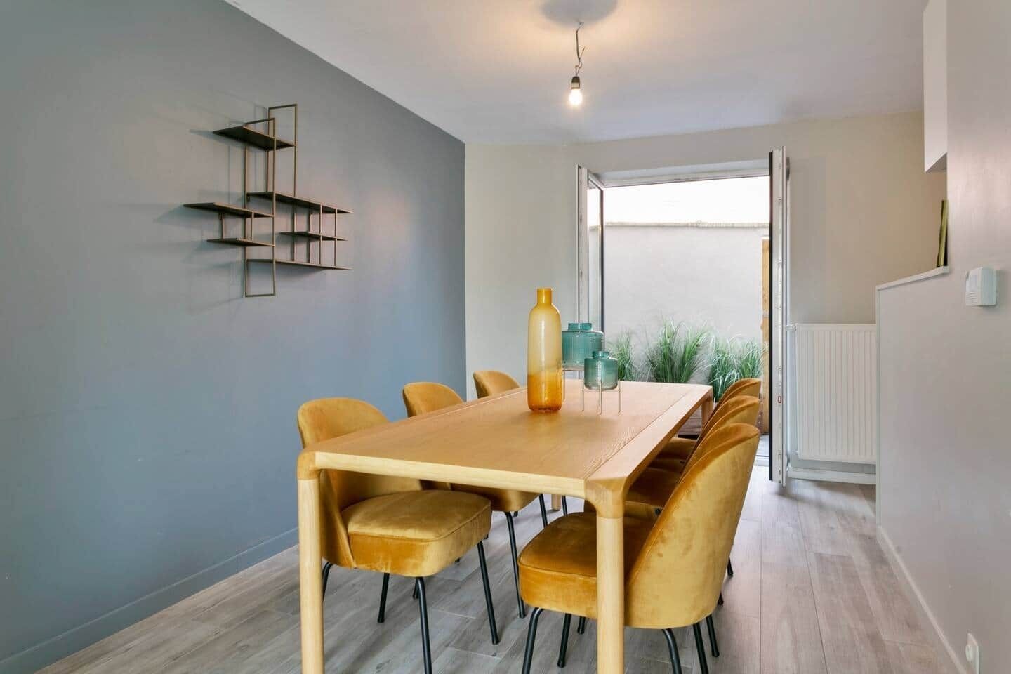 A dining area with a wooden table and velvet chairs, a pendant light, and a view through a large window.