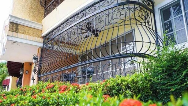 A building with a curved balcony with wrought iron railings, surrounded by well-maintained greenery and red flowers.