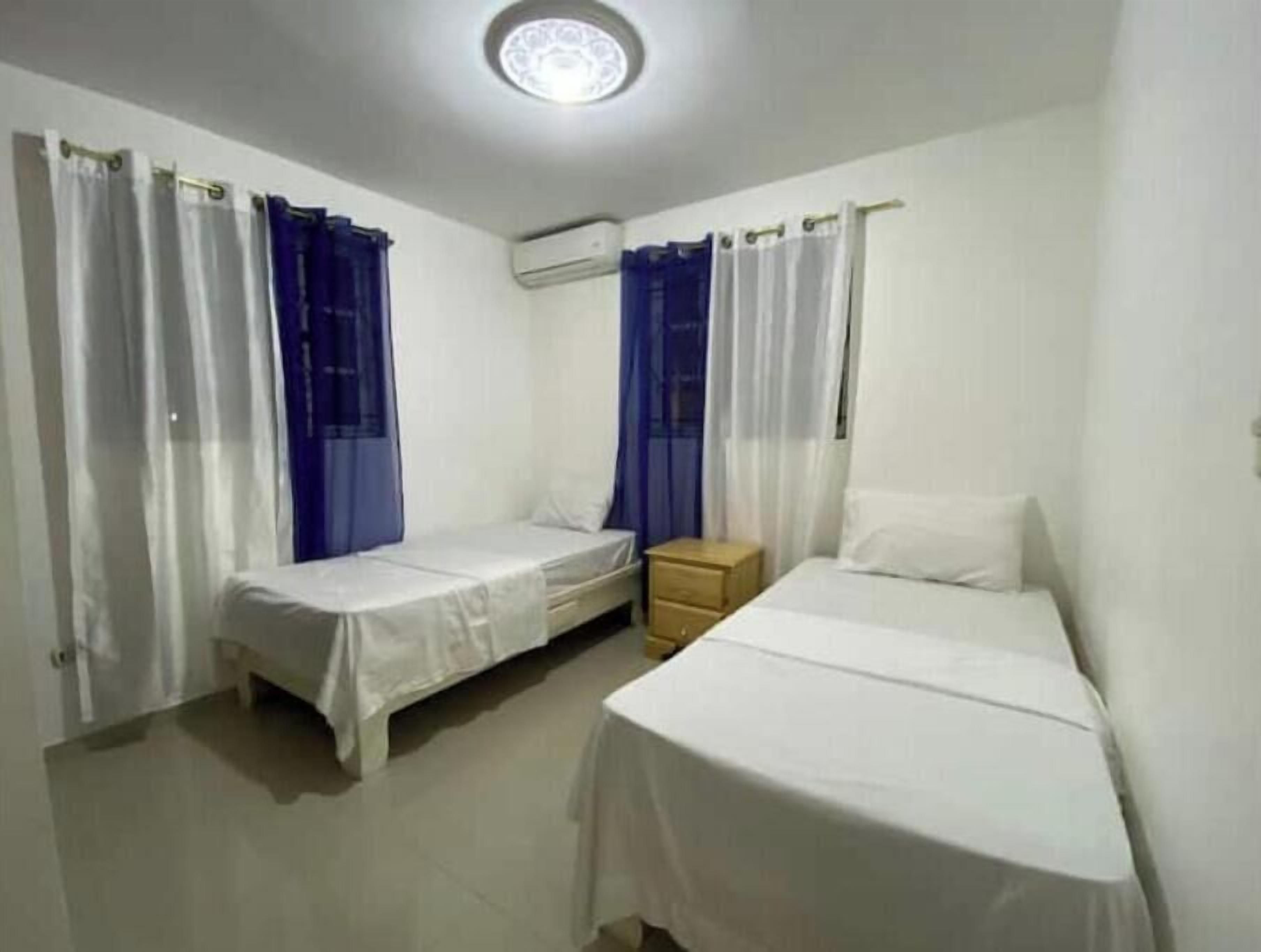 Una habitación pequeña y limpia con dos camas individuales, una mesita de noche de madera y un equipo de aire acondicionado en la pared.
