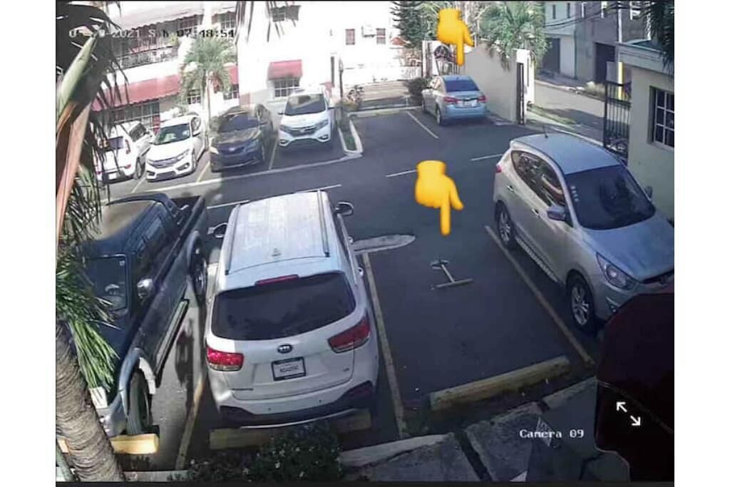 Parkkipaikalla on useita parkkeerattuja autoja, taustalla näkyy punaisella katoksella varustettu rakennus, ja maassa on kamera, jossa on merkintä ”Canera 09”.