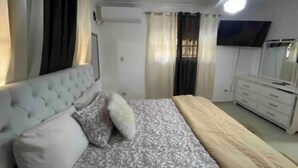A bedroom with a tufted headboard, a bed with a floral bedspread, a bedside table, a dresser, a ceiling fan, and a television on the wall. - Santo Domingo huge furnished longterm available apt (Santo Domingo norte)
