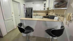 A compact kitchen with white cabinets, a central island, two black bar stools, a refrigerator, a stove, and a phone on the wall. - Santo Domingo huge furnished longterm available apt (Santo Domingo norte)