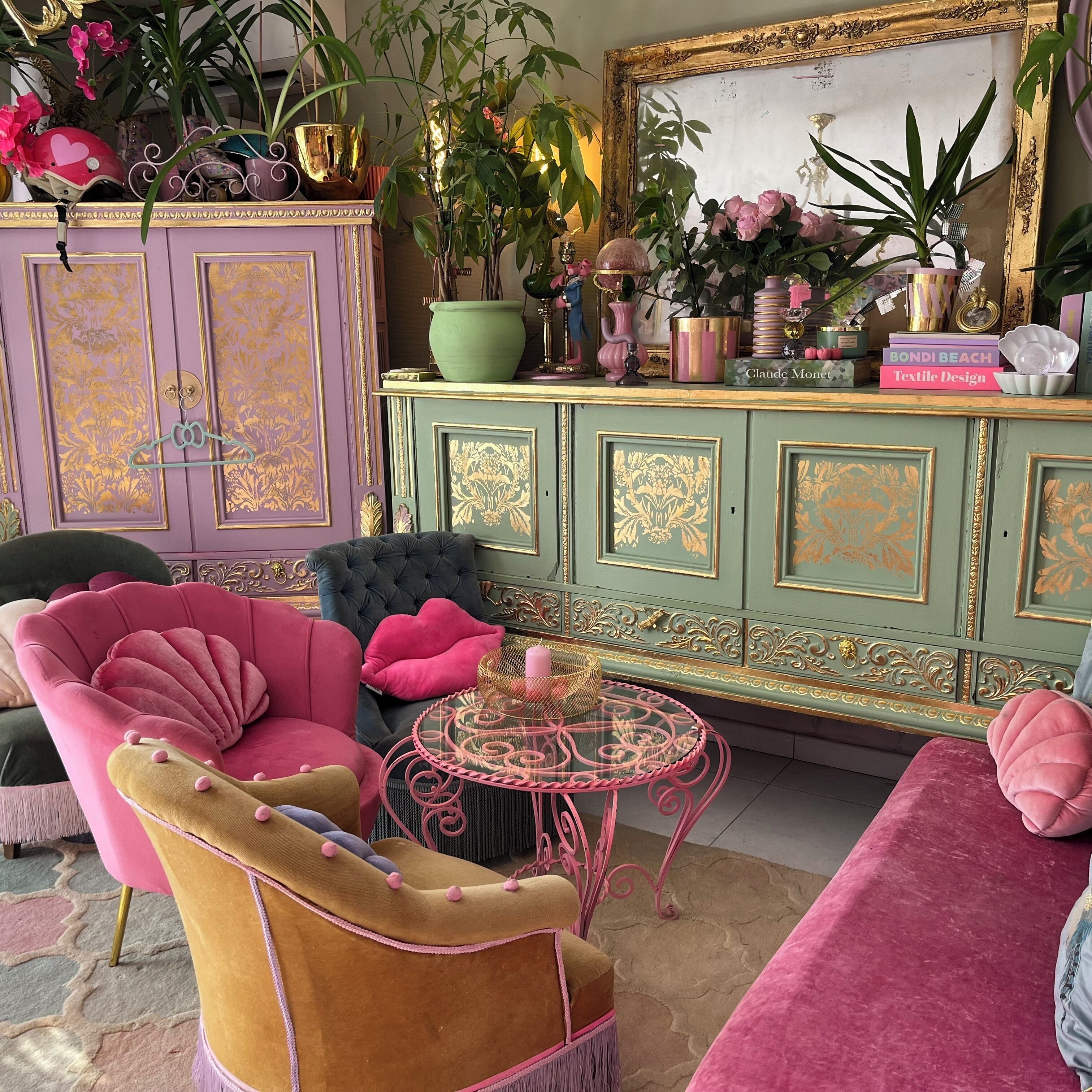 Een kamer met een roze fauteuil, een groene kast met gouden accenten, en een spiegel met een gouden lijst.