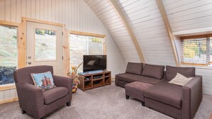 A cozy living room with a wooden ceiling, two sofas, a TV stand, and large windows offering a view of the outdoors.