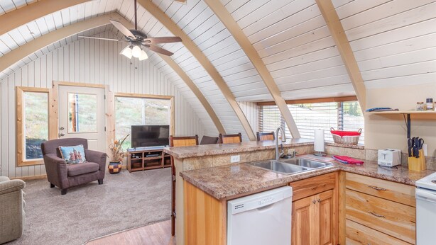A spacious living area with a kitchen, wooden ceiling beams, a ceiling fan, and a television.