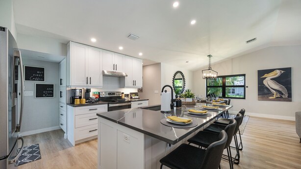 A modern kitchen with white cabinets, a central island with bar stools, a large window, and a painting of a pelican.