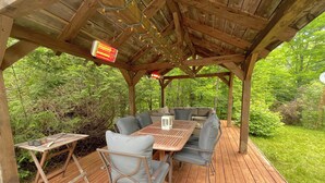 Une terrasse en bois dotée d’un espace salon abrité, comprenant une table et des chaises, le tout entouré d’une végétation luxuriante.