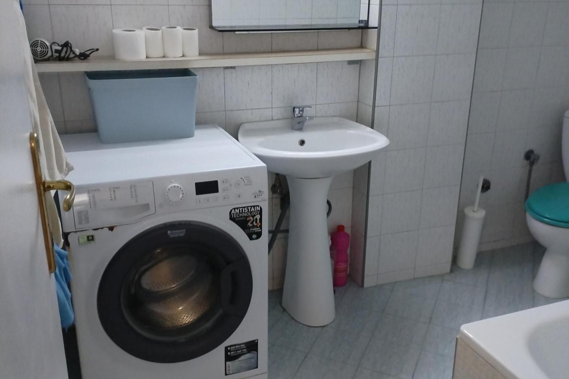 A compact bathroom with a front-loading washing machine, a pedestal sink, and a toilet with a green seat.