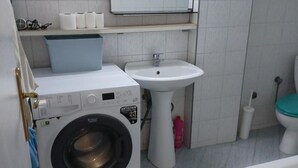 Un baño pequeño con lavadora de carga frontal, lavamanos pedestal y un inodoro con asiento verde.