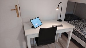Un escritorio pequeño y blanco con una tableta y un control remoto, una silla negra y una cama con ropa de cama estampada.