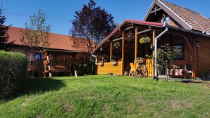 A wooden cabin with a red-tiled roof, a porch with hanging flower baskets, and a wooden swing set. - Casuta cu Brazi (Seusa)