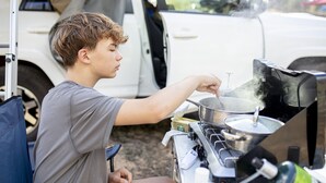 Eine Person kocht draußen mit einem Campingkocher, einem Topf und einer Ölflasche.