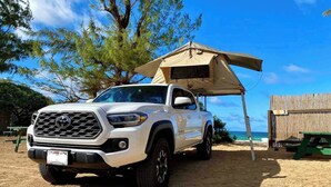 Una camioneta pickup blanca estacionada en una playa arenosa, con un toldo y una mesa de picnic cerca.