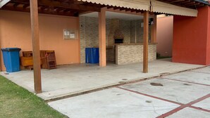 A covered patio area with a brick fireplace, wooden furniture, and a blue trash bin. - Golden Lake (Arraial do Cabo)