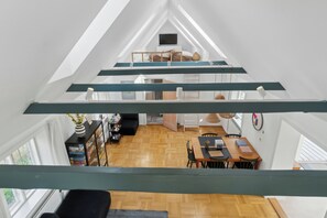 A loft-style room with a wooden floor, a dining area with a table and chairs, a TV on a shelf, and a staircase leading to an upper level.