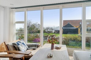 A room with a large window, a wooden table, a cushioned chair, and a vase with flowers.