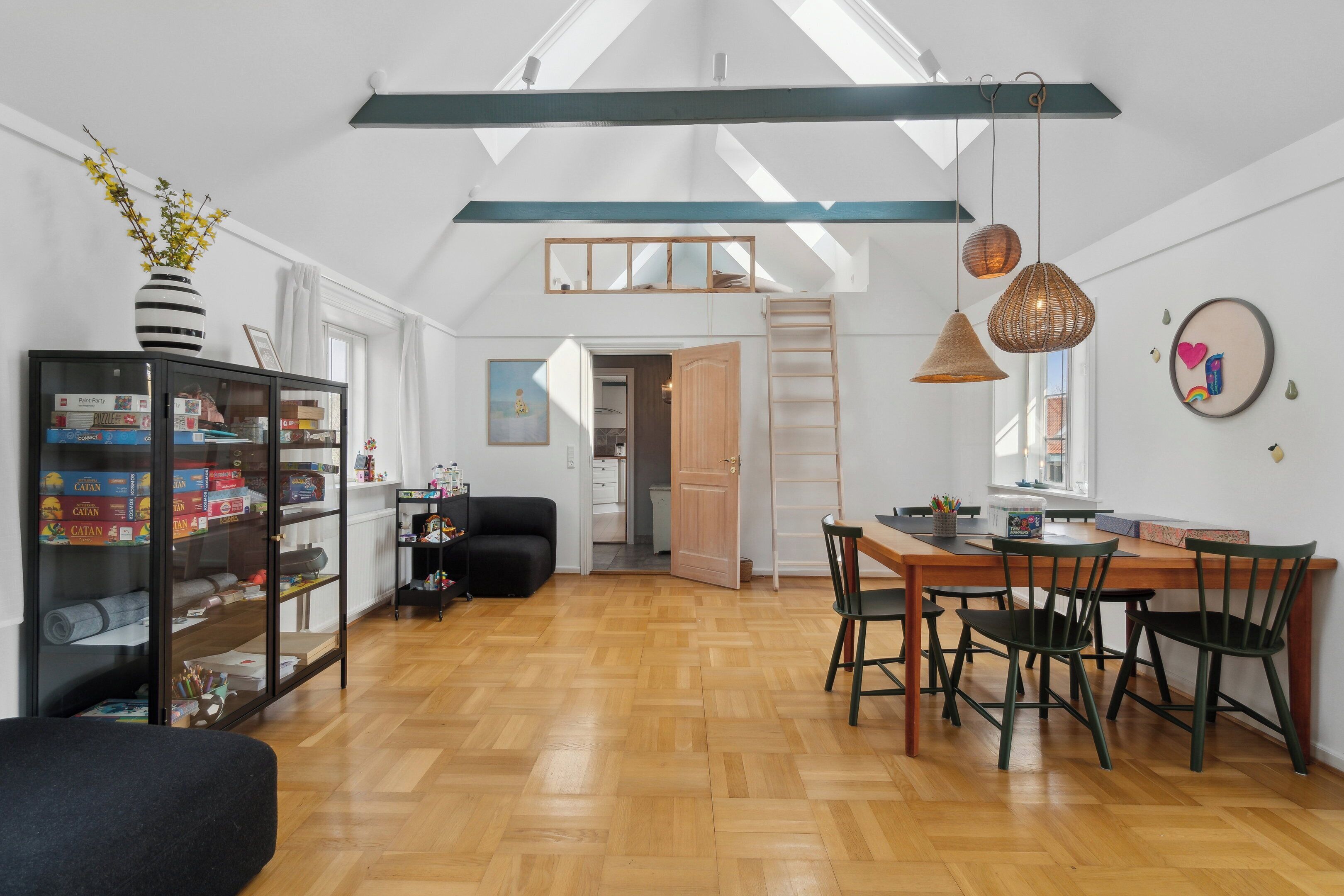 A spacious loft with a high ceiling, wooden flooring, a dining area with a table and chairs, a black sofa, and a bookshelf filled with board games.