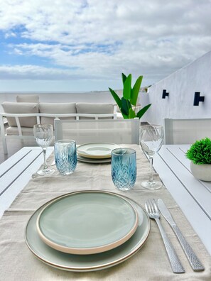 A dining table set with plates, glasses, and cutlery, overlooking a coastal view with a white outdoor seating area.